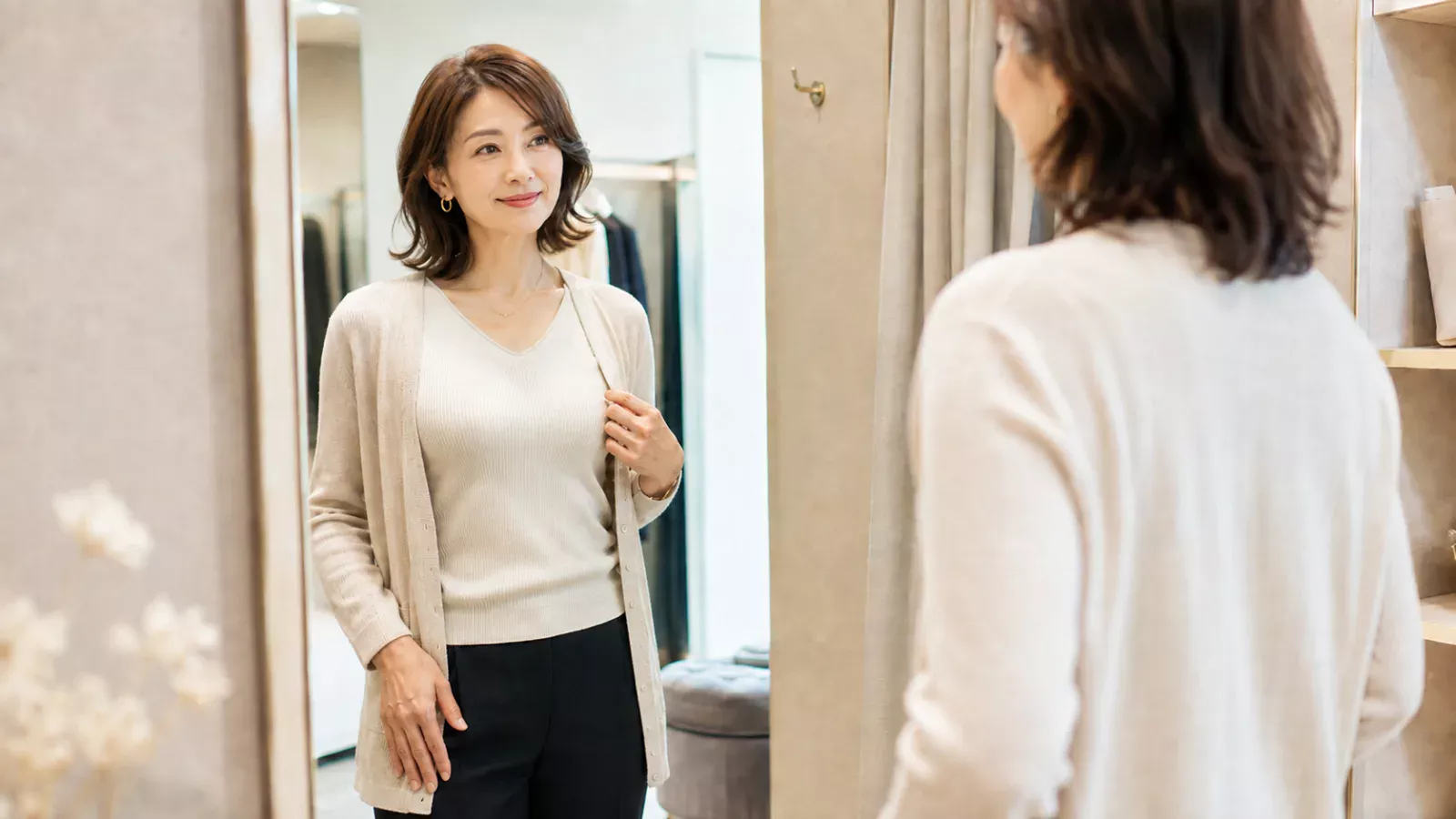 明るく上品なフィッティングルームで、40代女性が洋服を着た状態で鏡を見ながら胸元のシルエットを確認している様子。
